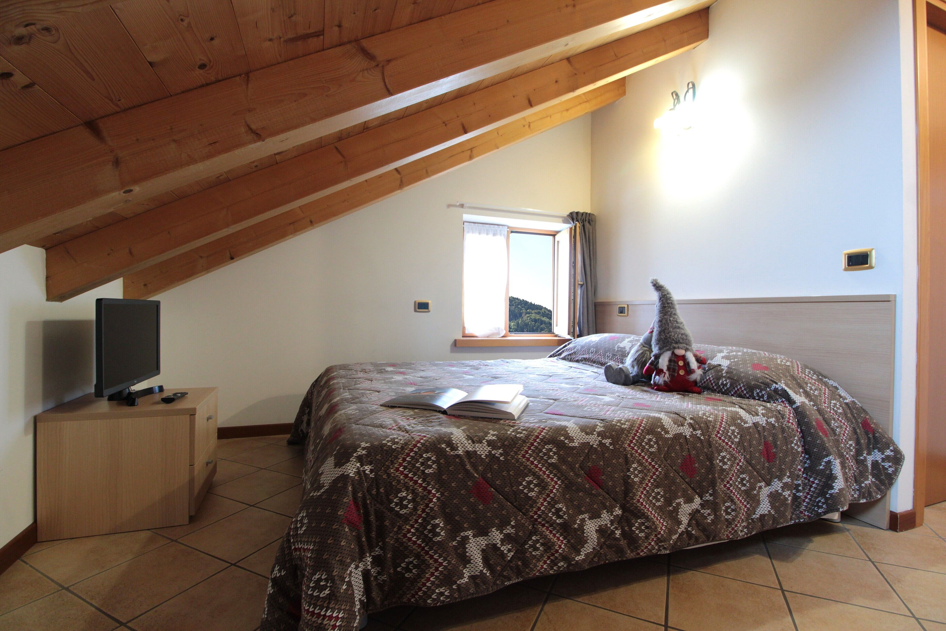 Un dormitorio con techo inclinado, una cama con colcha estampada, un libro, un televisor sobre una mesita de madera y una ventana con cortinas.