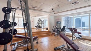A gym with weightlifting equipment, cardio machines, and large windows with a city view. - Stayshort - Stylish 1BR Apartment With Stunning Lake Views (Dubai)
