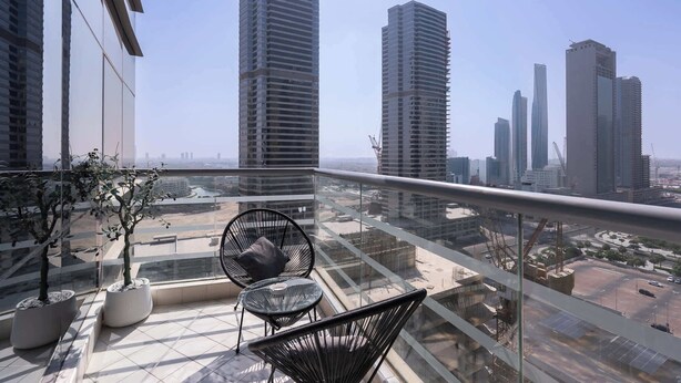 A balcony with a view of high-rise buildings, a glass railing, and two potted plants. - Stayshort - Stylish 1BR Apartment With Stunning Lake Views (Dubai)