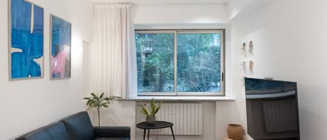 A modern living room with a black leather sofa, a small round table with a plant, a large window with a view of trees, and abstract art on the wall.