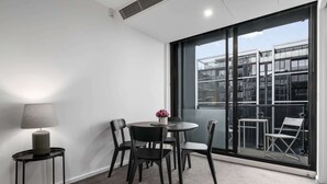A modern room with a round table, four chairs, a lamp, and a large window with a view of buildings. - City Stay Steps From Yarra River Walks (Melbourne)