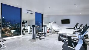 A modern gym with large windows, exercise equipment, and a city view. - City Stay Steps From Yarra River Walks (Melbourne)