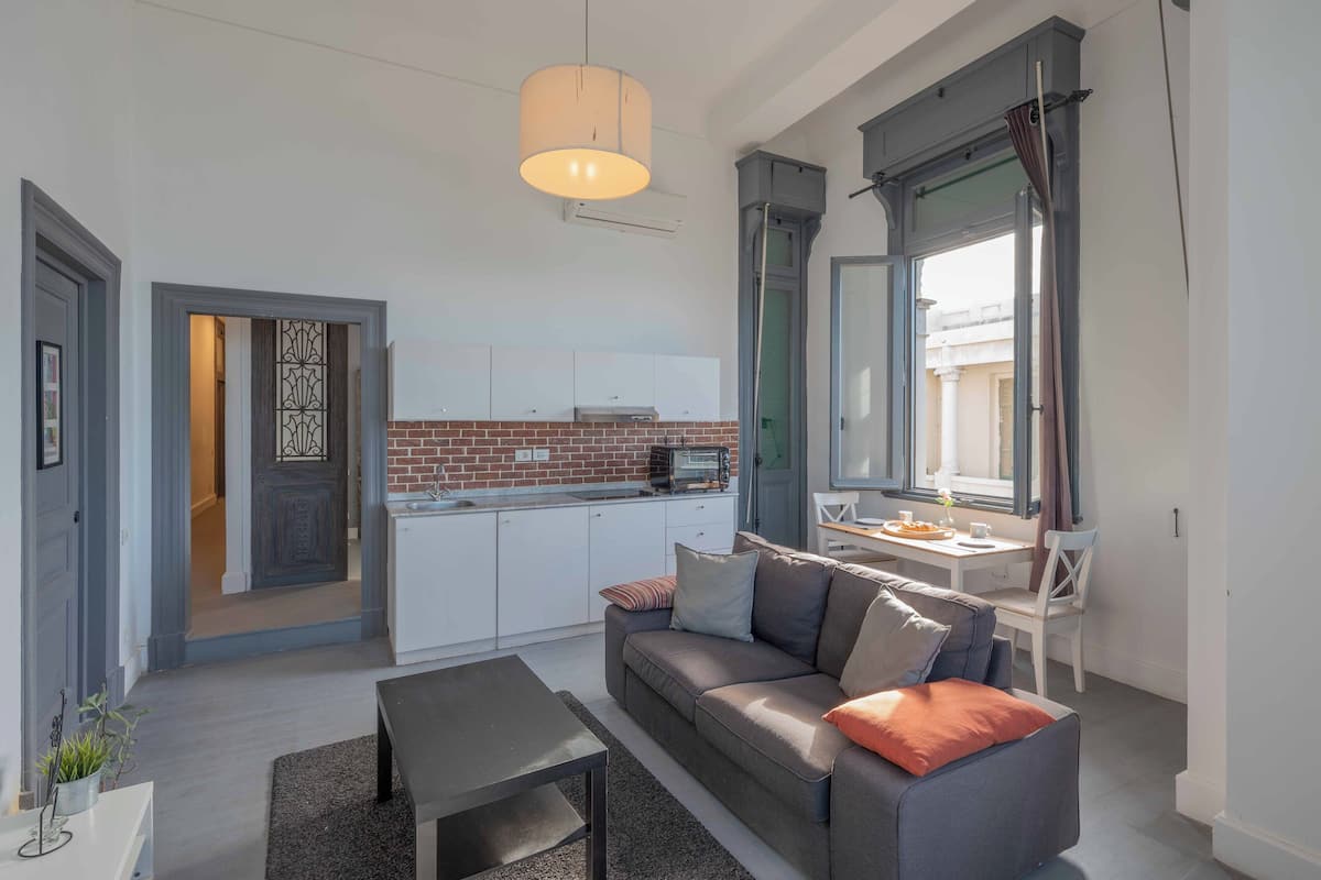 Een knus woongedeelte met een kitchenette, een bank, een eettafel en een raam met gordijnen.