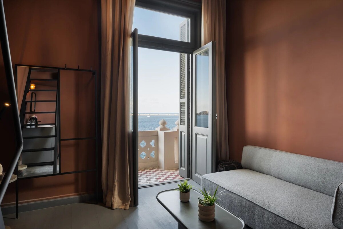 Un balcón con vista al mar, un sofá, una pequeña mesa con plantas y una escalera.