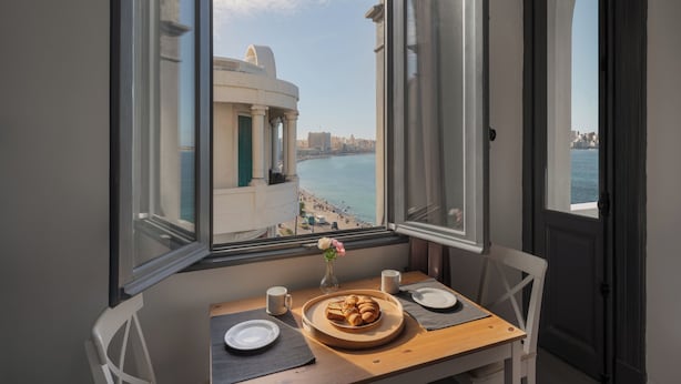A room with a table set for breakfast, a window with a view of a beach and cityscape, and a door. - Grey l studio apartments Corniche Alexandria Little Venice (Alexandria)