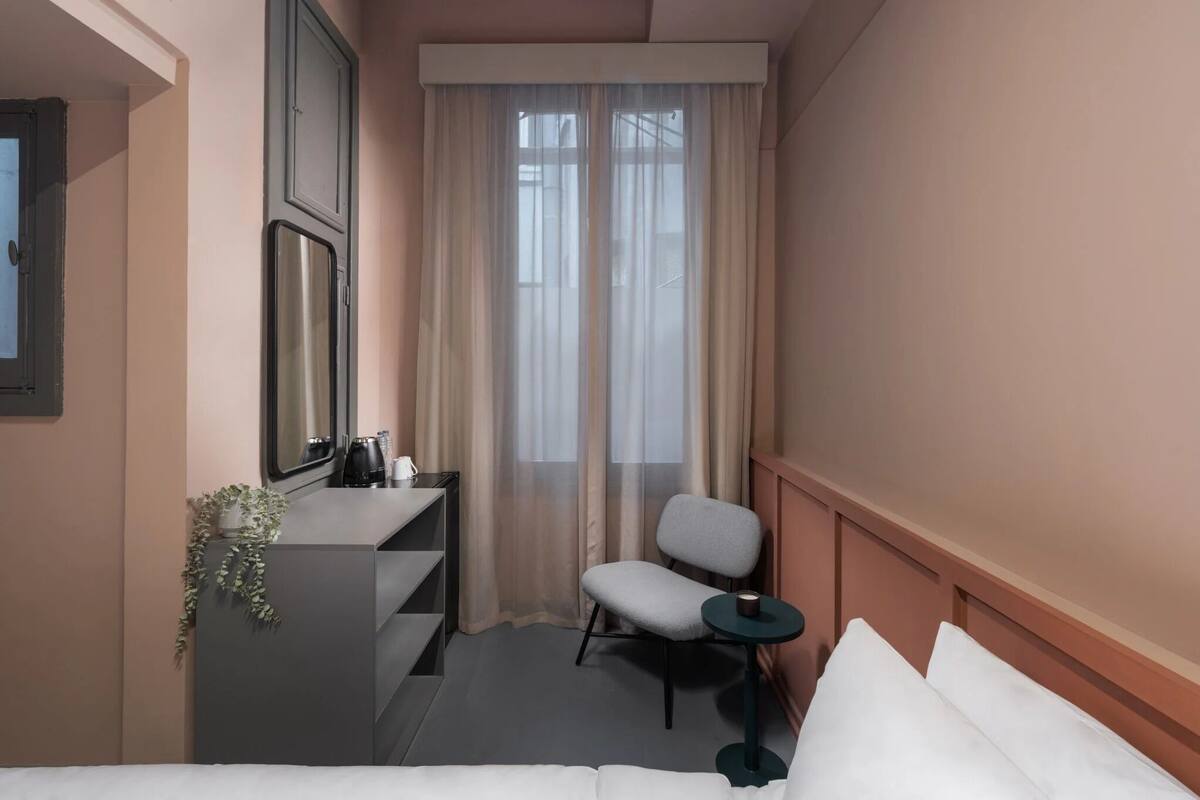 Een hotelkamer met een bed, een bureau met stoel, een tafeltje, een plant en een raam met gordijnen.