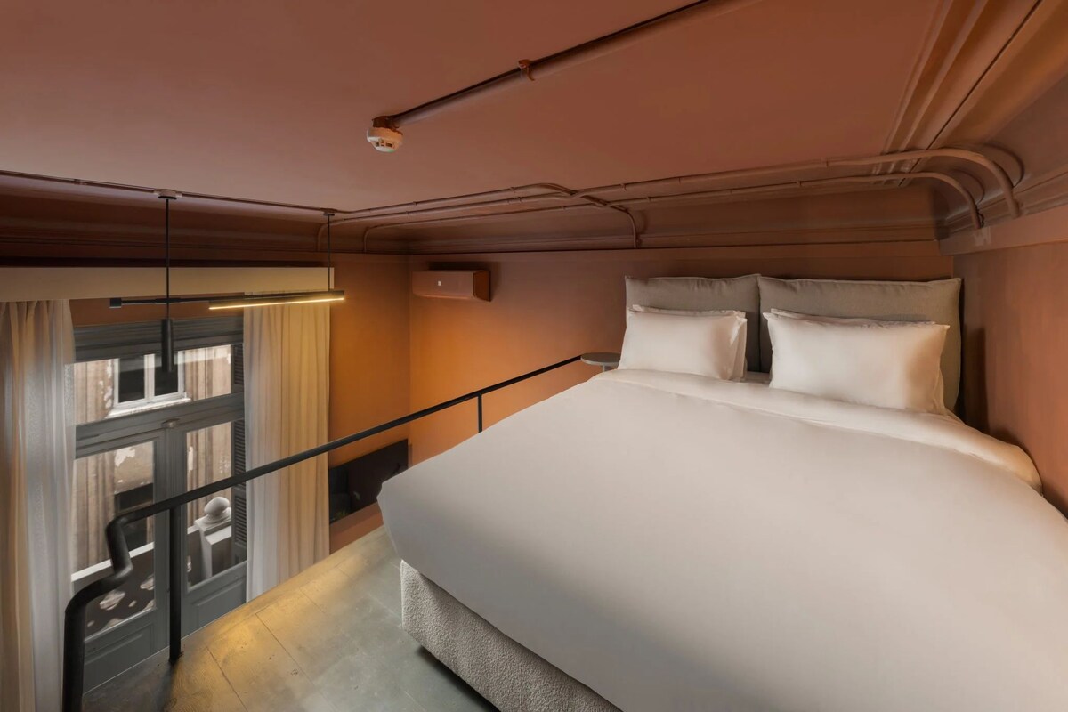 Un dormitorio con cama, ventana con cortinas, barandilla y una lámpara en el techo.