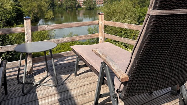 A wooden deck with a table and chairs overlooking a lake, surrounded by greenery and buildings in the background. - House Königsee *garden view (Bad Oldesloe)