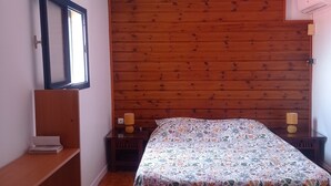 Ein Schlafzimmer mit Holzbalkendecke, einem Bett mit floralem Überwurf, einem Nachttisch mit Lampe und einem Regal mit einem Buch.