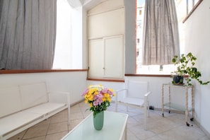 A room with a glass table, a vase of flowers, a white bench, a white chair, and a small table with a potted plant.