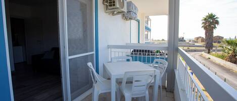A balcony with a white table and chairs, an air conditioning unit, and a view of the street and palm trees.