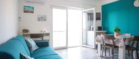 En moderne stue med en turkis sofa, et spisebord med stole og et køkkenområde, der kan ses igennem en åben dør.