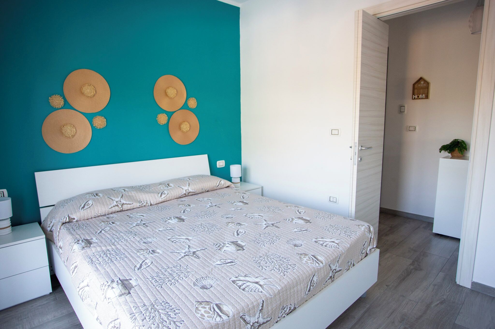 Un dormitorio con una pared de acento color turquesa, una cama con colcha de estampado floral y una mesita de noche blanca con lámpara.