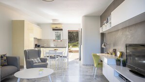 Un salon moderne avec un canapé, un fauteuil et une table basse. Il y a un coin cuisine avec des placards et une table à manger. Un téléviseur est fixé au mur, et il y a des plantes sur le comptoir.