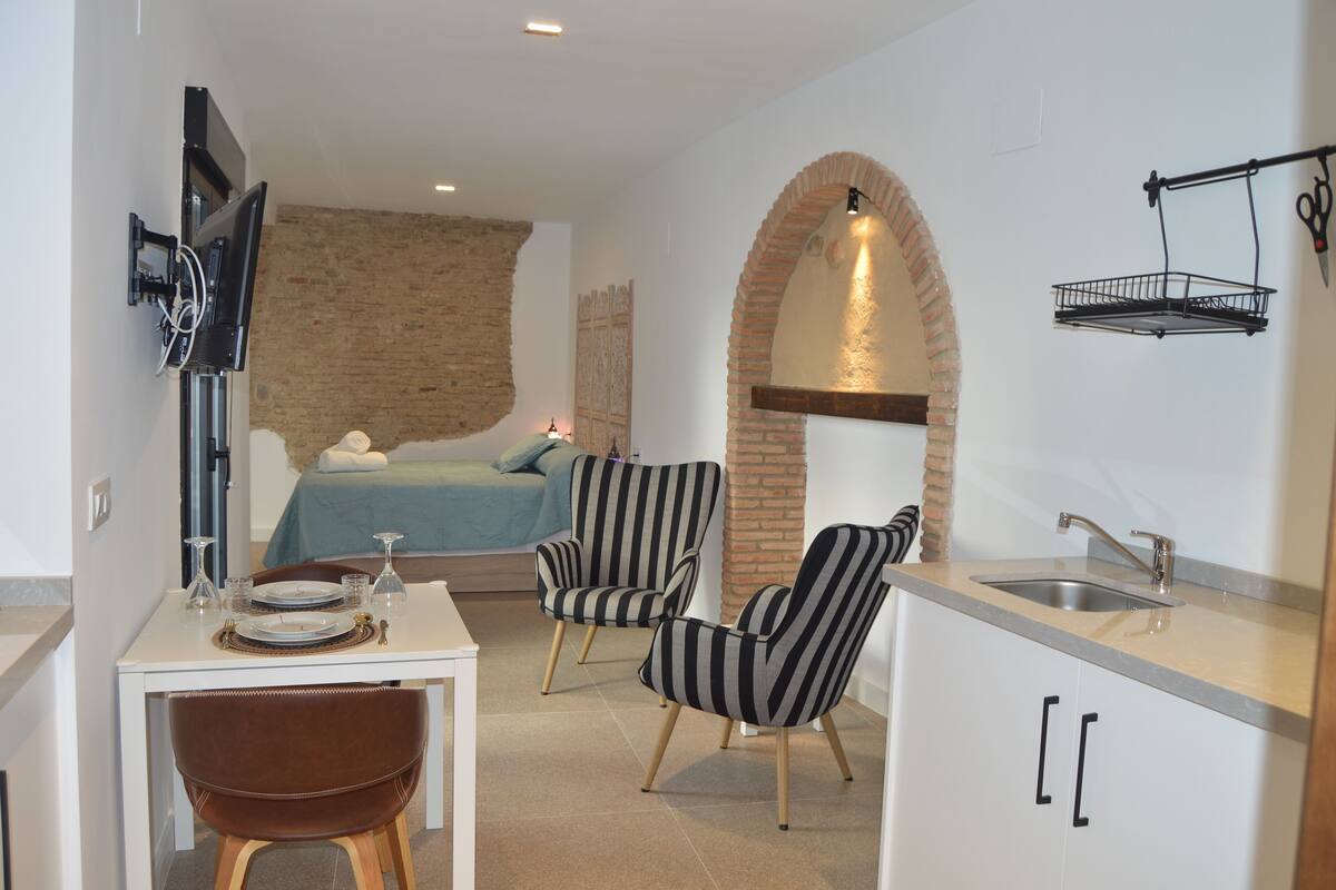 A compact room with a bed, a dining table with chairs, a kitchen area with a sink, and a TV mounted on the wall.