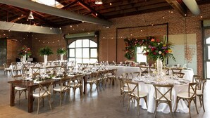 Une vaste salle dotée de hauts plafonds et de murs en briques apparentes, aménagée avec des tables rondes et des chaises en bois, idéale pour l’organisation d’événements.
