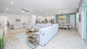 A spacious living area with a large white sectional sofa, a dining table with chairs, and a kitchen in the background. - Upstay - Magic Mansion w Pool-spa-cinema-games (Kissimmee)