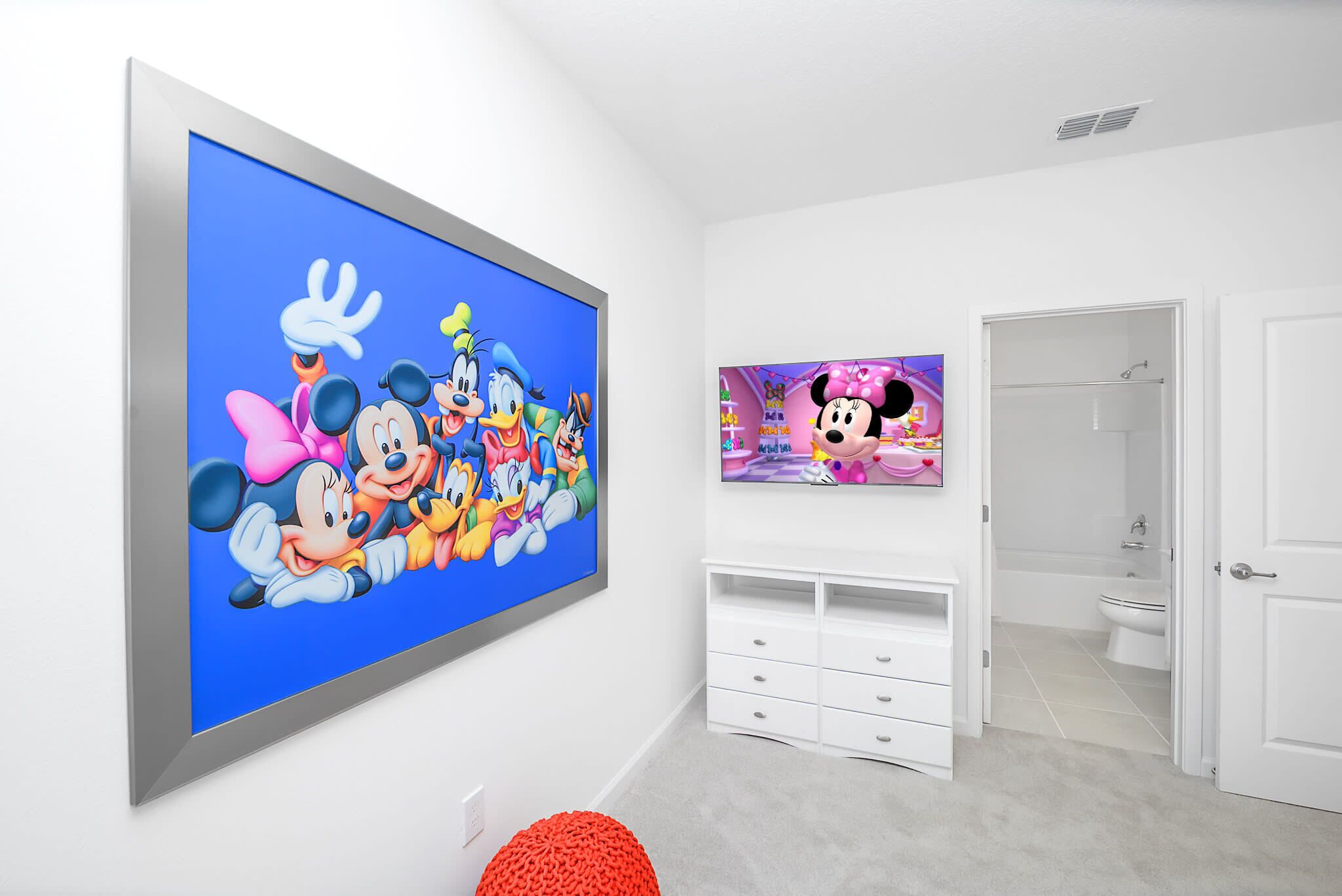 Une pièce dotée d’un grand téléviseur à écran plat diffusant des dessins animés, d’une commode blanche et d’une salle de bain visible à travers une porte ouverte.