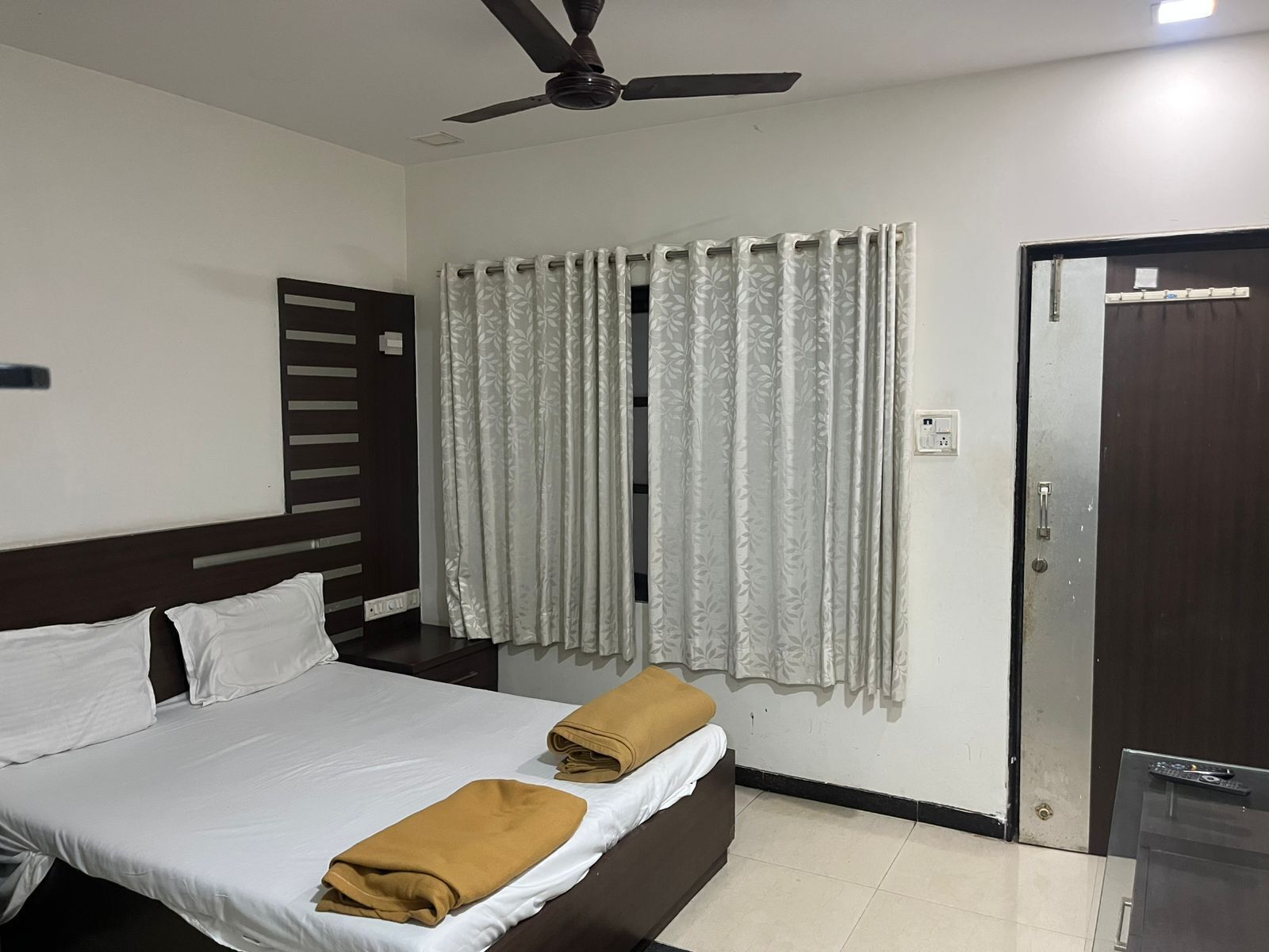 A hotel room with a bed, a ceiling fan, a window with curtains, and a door.
