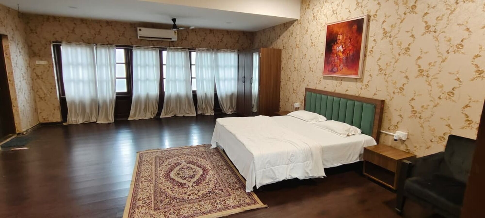 Un dormitorio amplio con una cama grande, un cabecero tapizado de color verde, una mesita de noche de madera y una alfombra estampada.