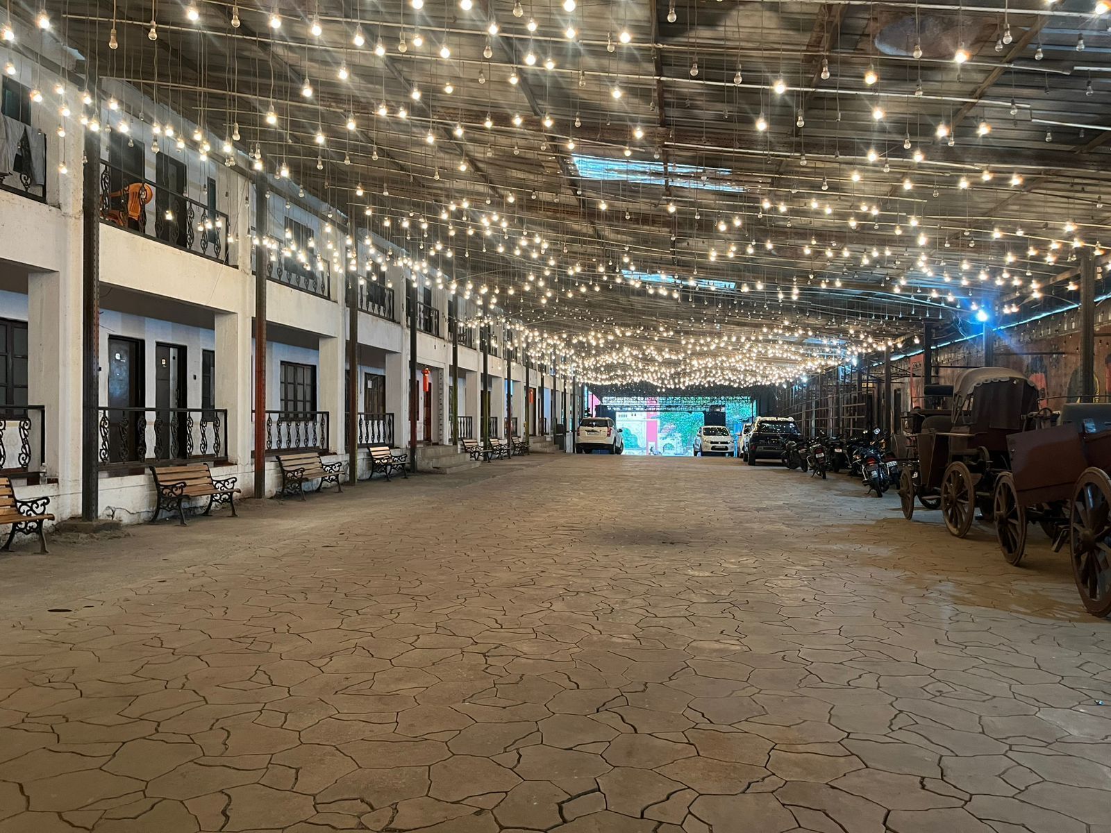 A spacious indoor area with a high ceiling adorned with numerous hanging lights, benches along the walls, and a variety of vehicles including cars and vintage carriages.