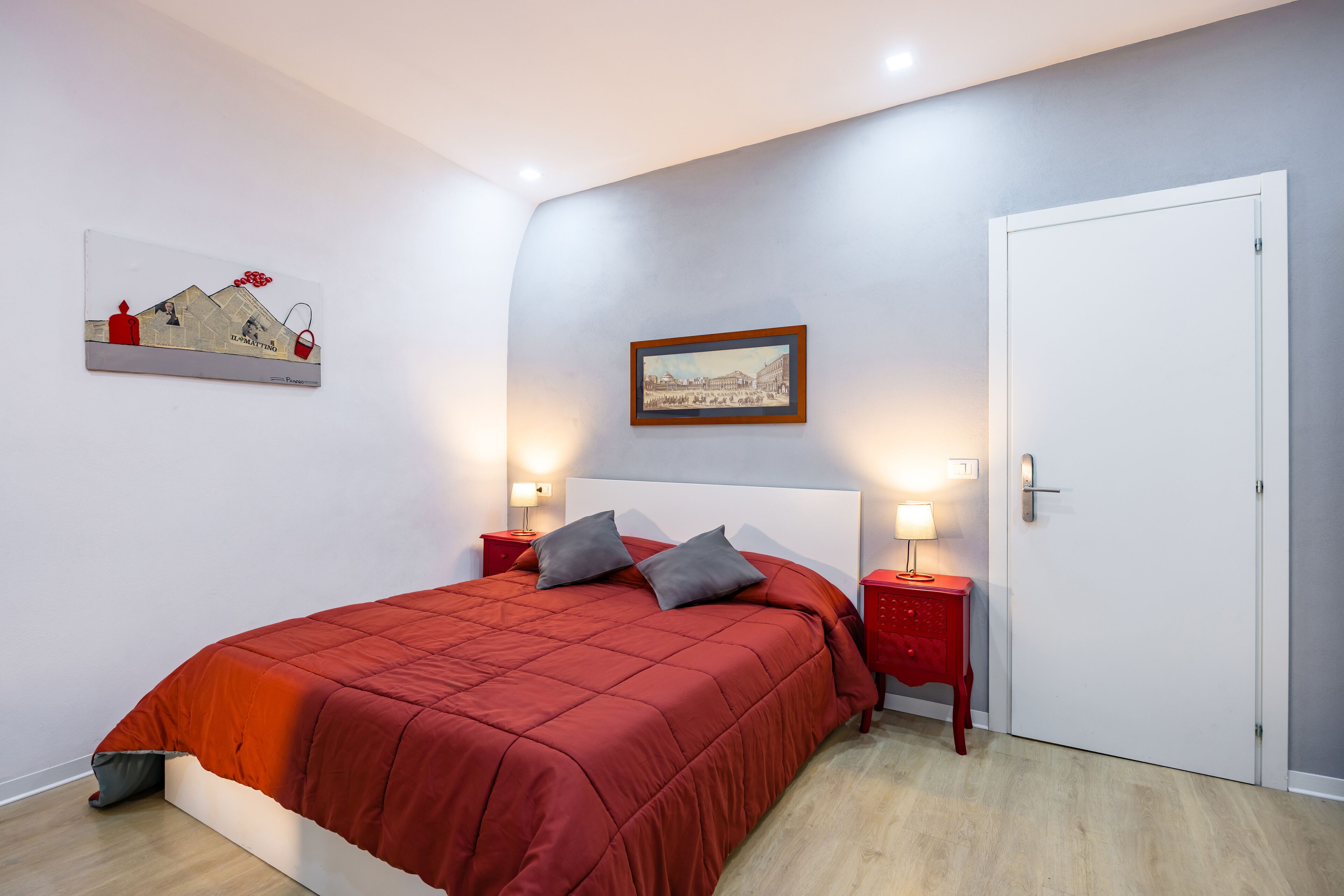 A bedroom with a red bedspread, two bedside tables with lamps, a door, and artwork on the walls.