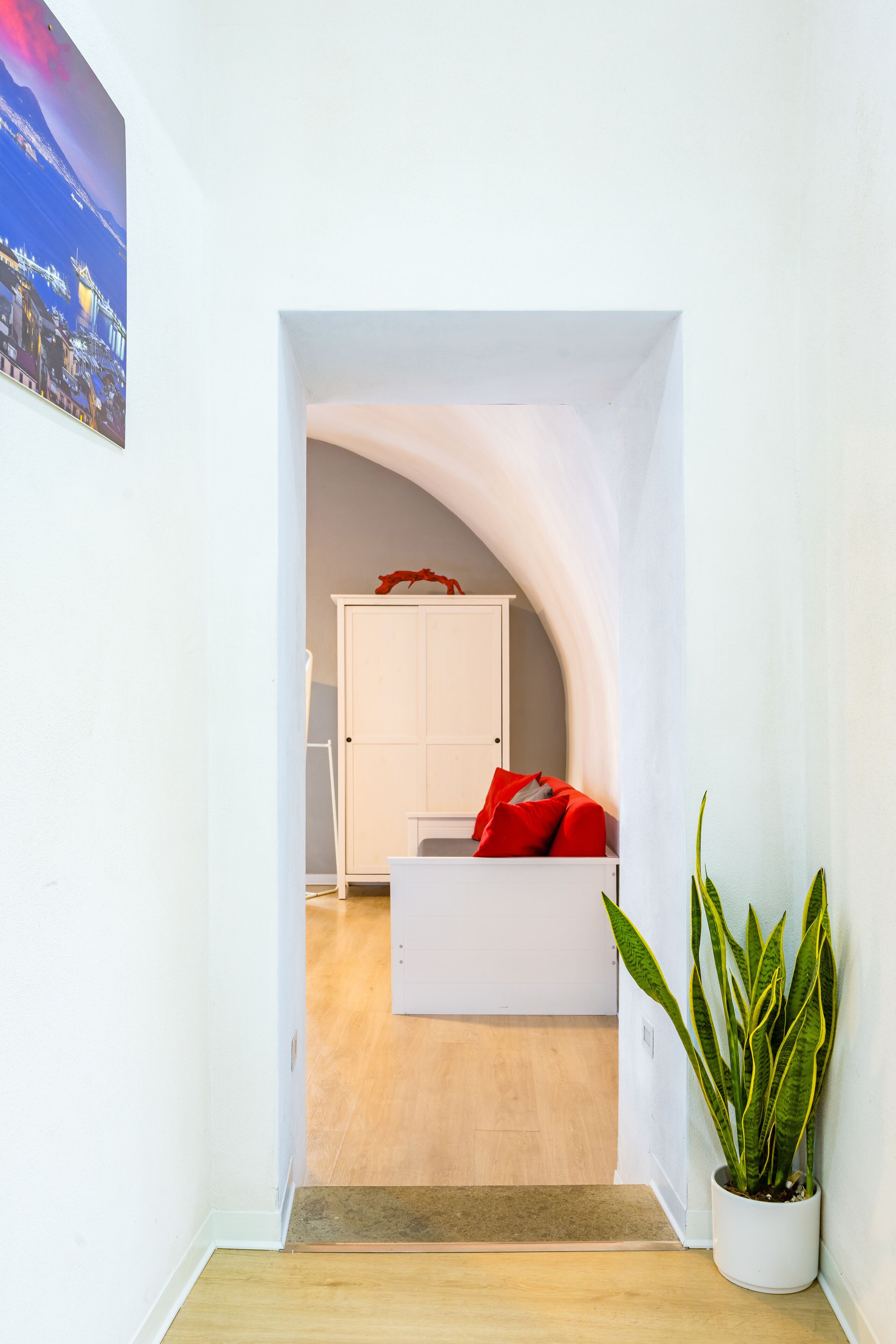 A hallway leading to a room with a white cabinet, a red pillow on a white sofa, and a potted plant.