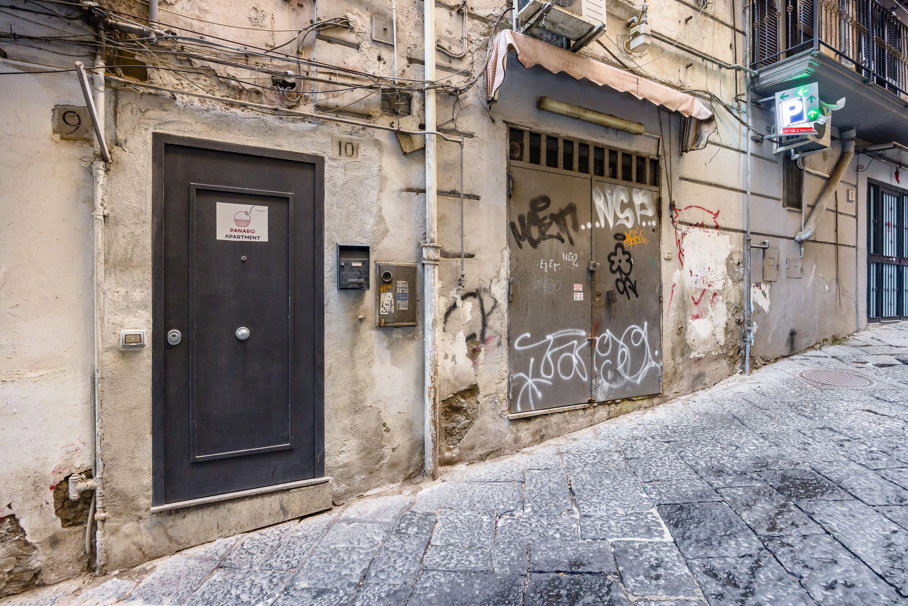 Un immeuble avec une porte fermée portant l’inscription « APPARTEMENT PANARO », une fenêtre recouverte de graffitis et un panneau de signalisation verte indiquant un espace de stationnement.