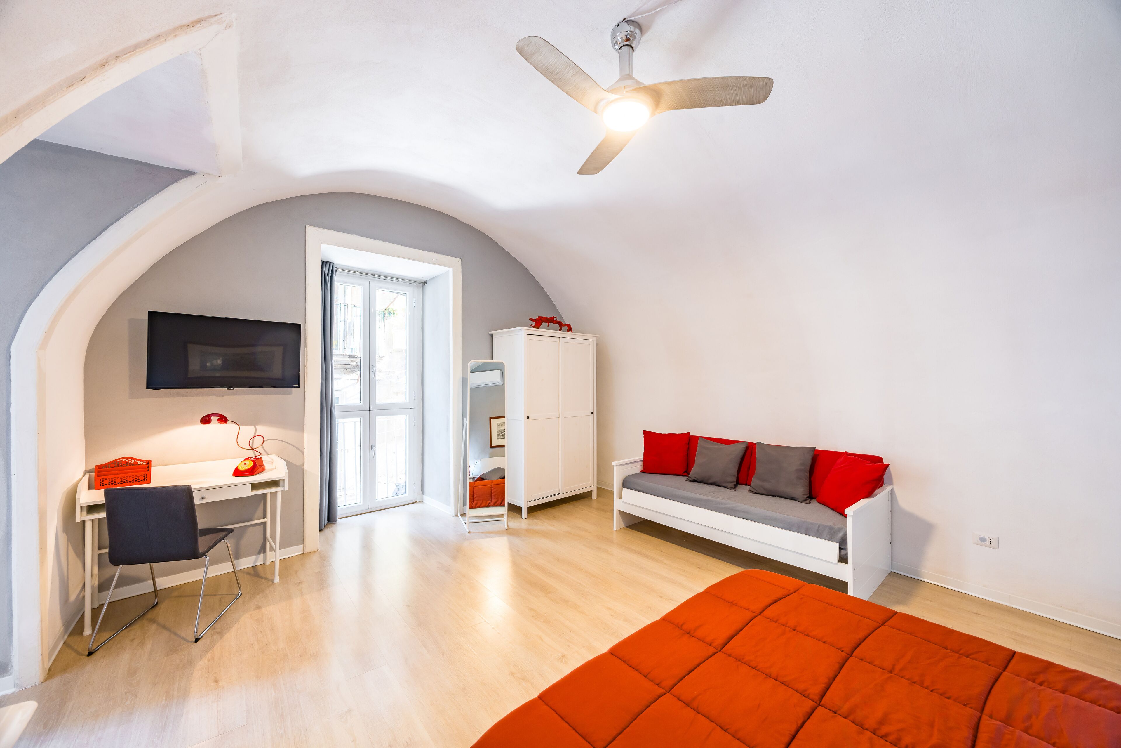 Ein modernes Schlafzimmer mit weißem Schreibtisch, schwarzem Sessel, weißem Schlafsofa mit roten und grauen Kissen und einem großen orangefarbenen Bett.
