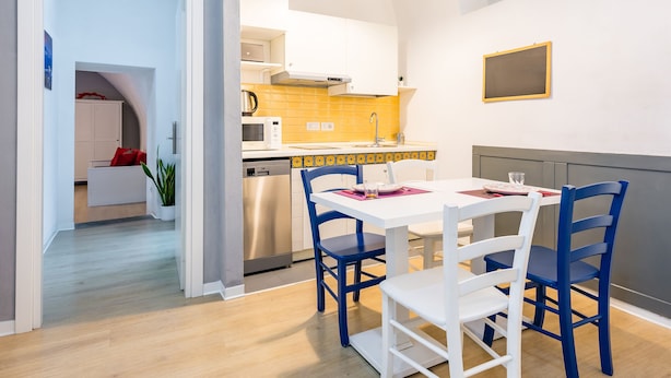 A modern kitchen with white cabinets, a stainless steel dishwasher, and a dining area with a white table and blue chairs. - O Panaro Lucky Flat by NapoliApartments (Naples)