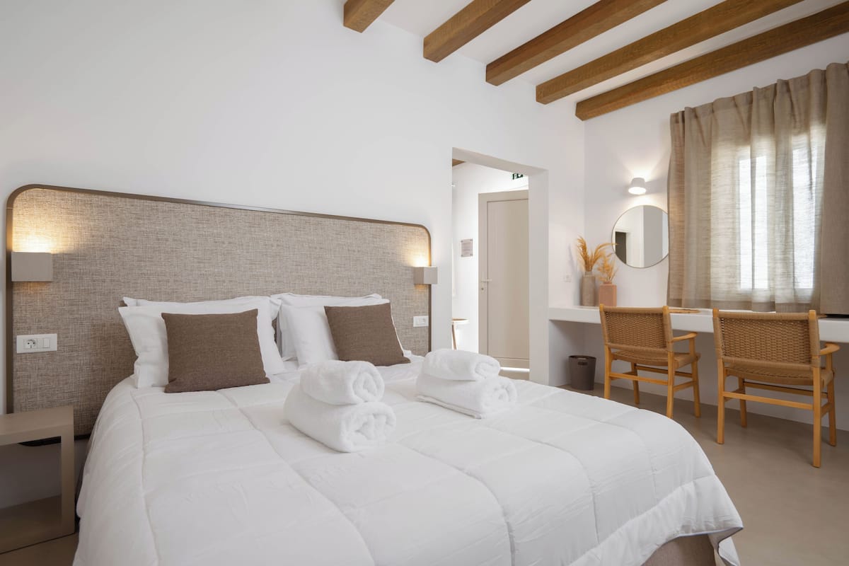 Una cama bien hecha con ropa blanca y almohadas marrones, un cabecero de madera, un escritorio con una silla y un espejo.