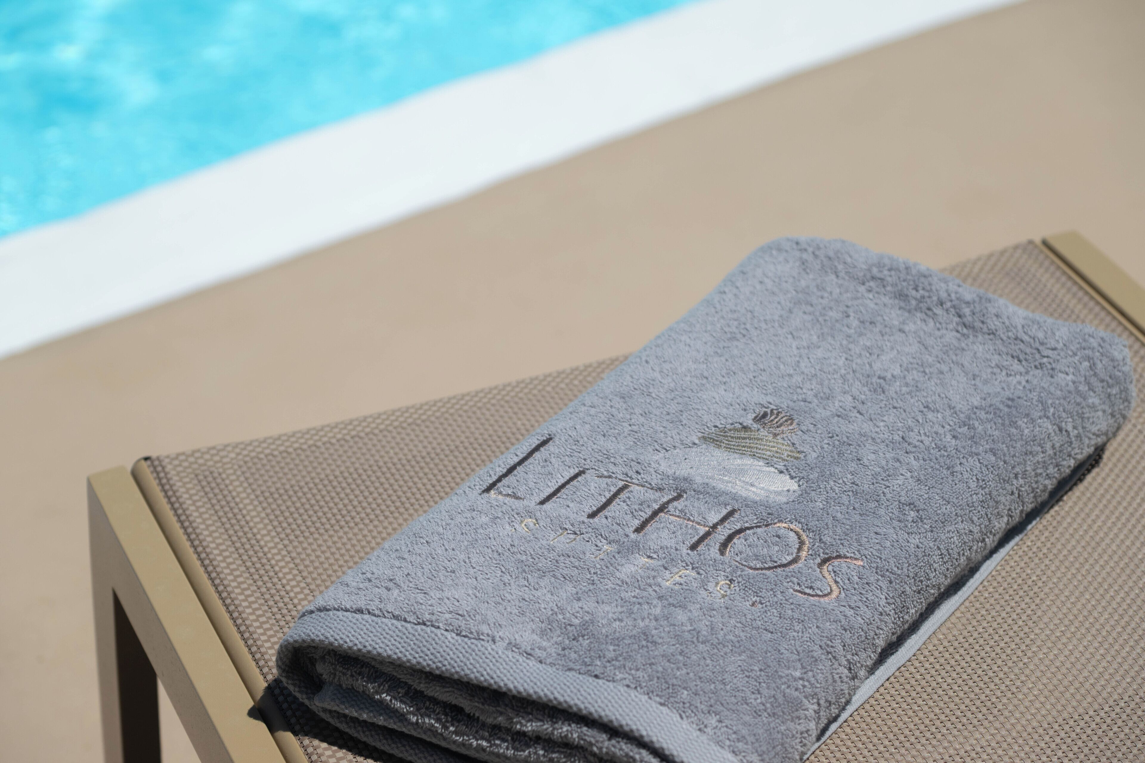 Una toalla doblada con el bordado 'LITHOS', sobre una silla junto a una piscina.