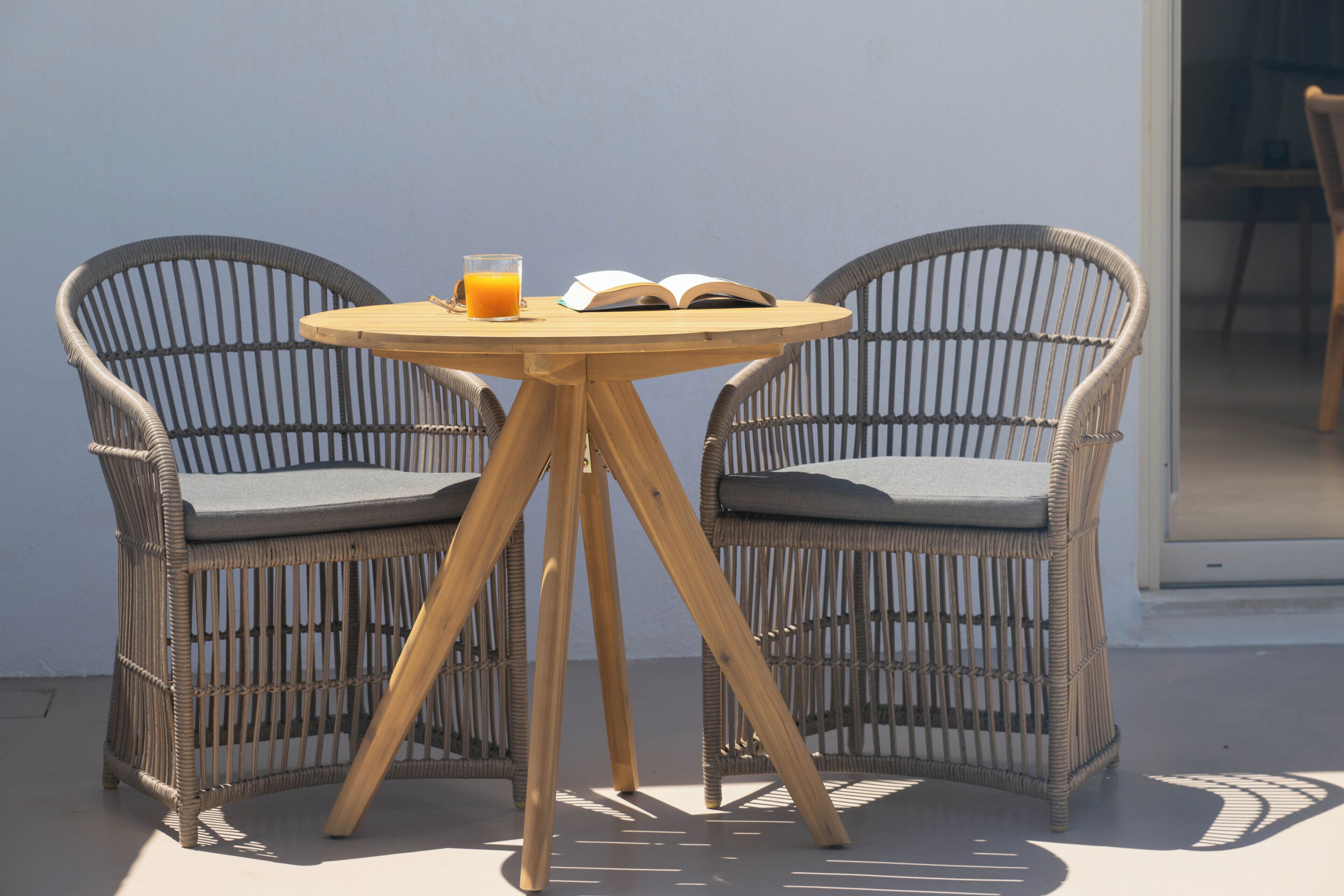 Un juego de silla y mesa de mimbre frente a una pared blanca, con un vaso de jugo de naranja y un libro abierto sobre la mesa.