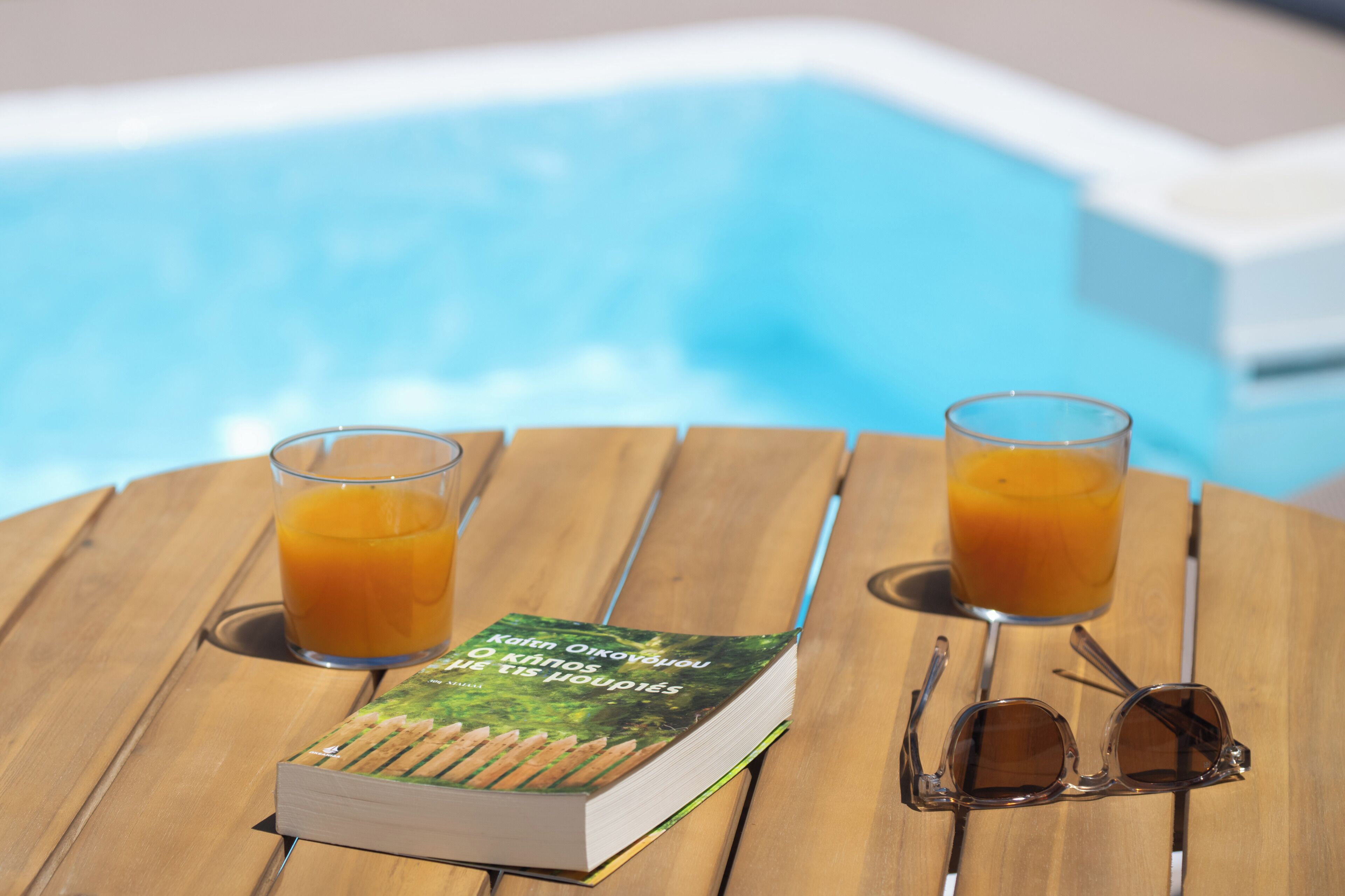 A book, two glasses of orange juice, and a pair of sunglasses on a wooden table by a pool.