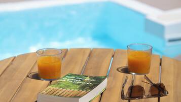A book, two glasses of orange juice, and a pair of sunglasses on a wooden table by a pool.
