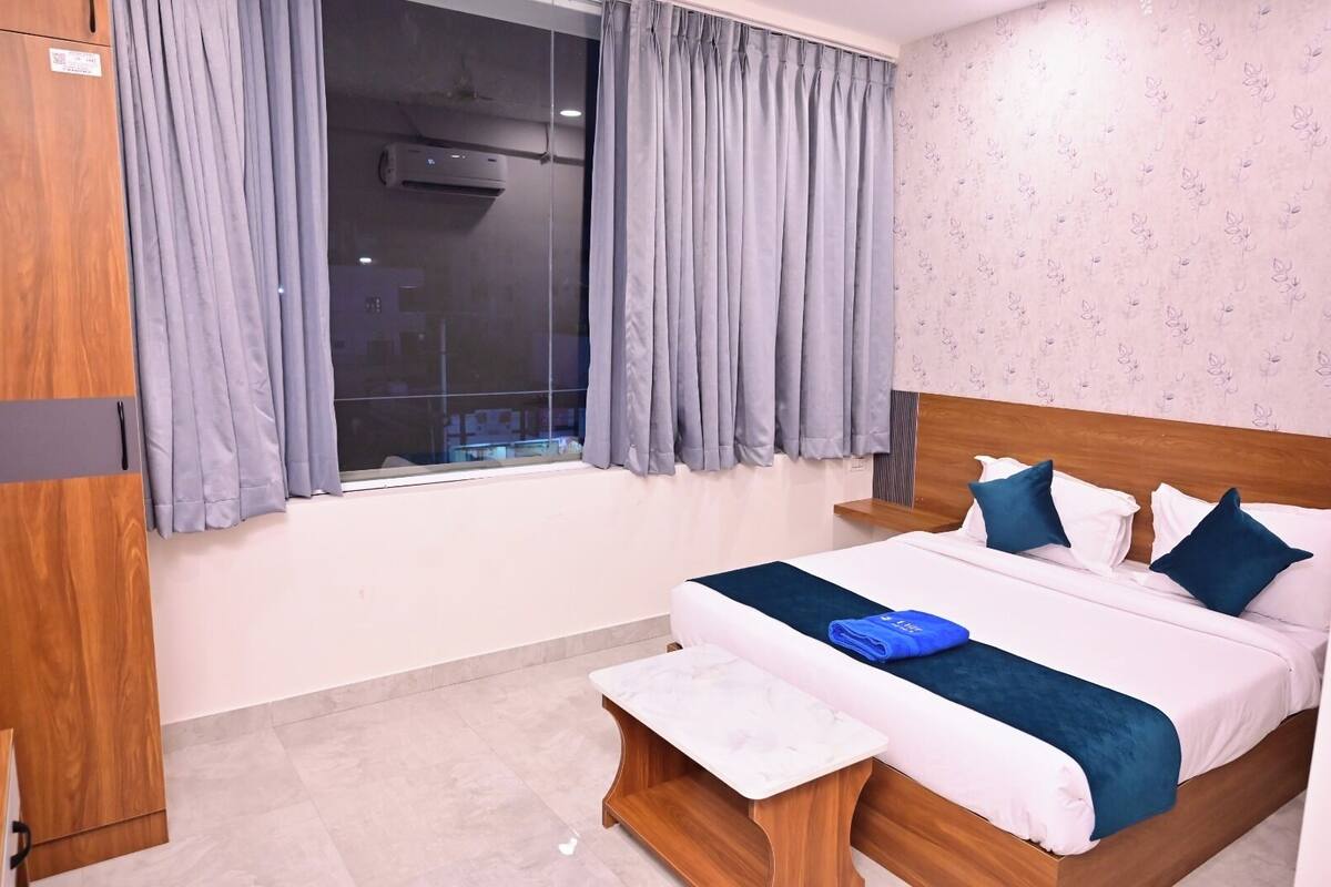 A hotel room with a bed, a wooden wardrobe, a window with curtains, and a wall with a floral pattern.