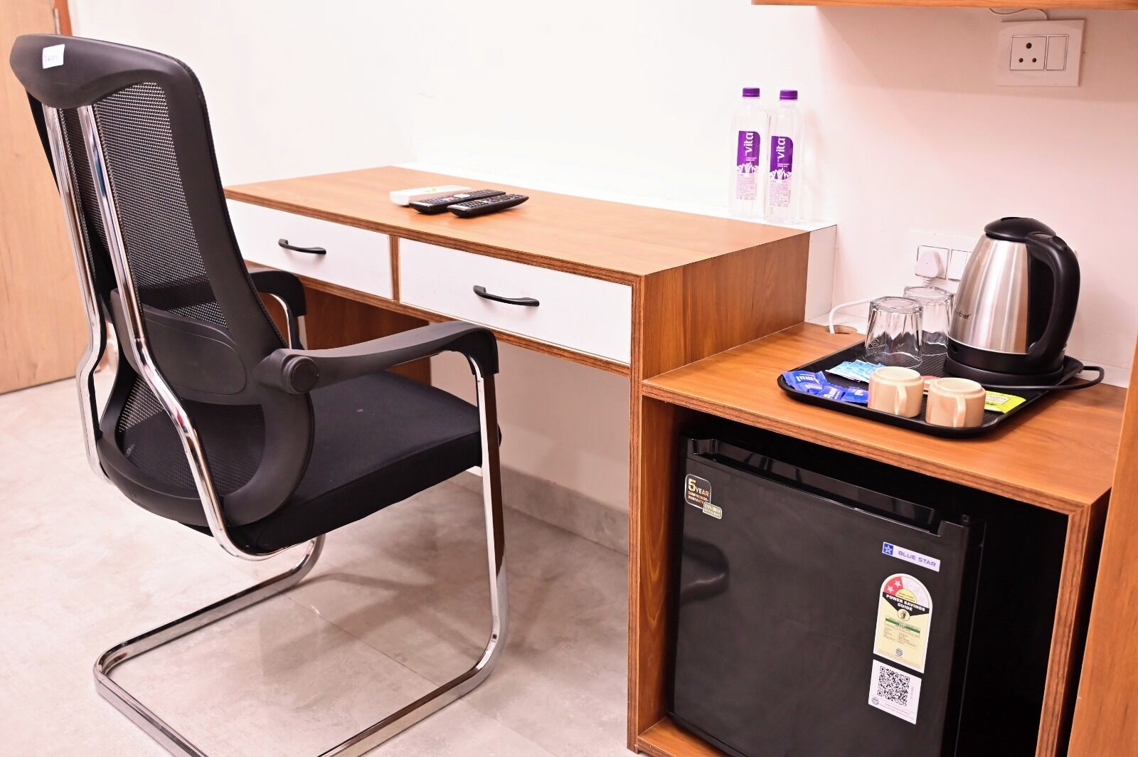 A compact hotel room with a single bed, a desk with a chair, a refrigerator, and a small kitchenette area.