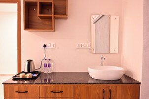 A modern bathroom with a wooden vanity, a white sink, and a mirror above it. There are two bottles of water on the vanity and a kettle on the countertop.