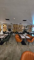 A modern restaurant with high ceilings, recessed lighting, and a patterned wall divider. Tables are set with menus and chairs are upholstered in green and orange.