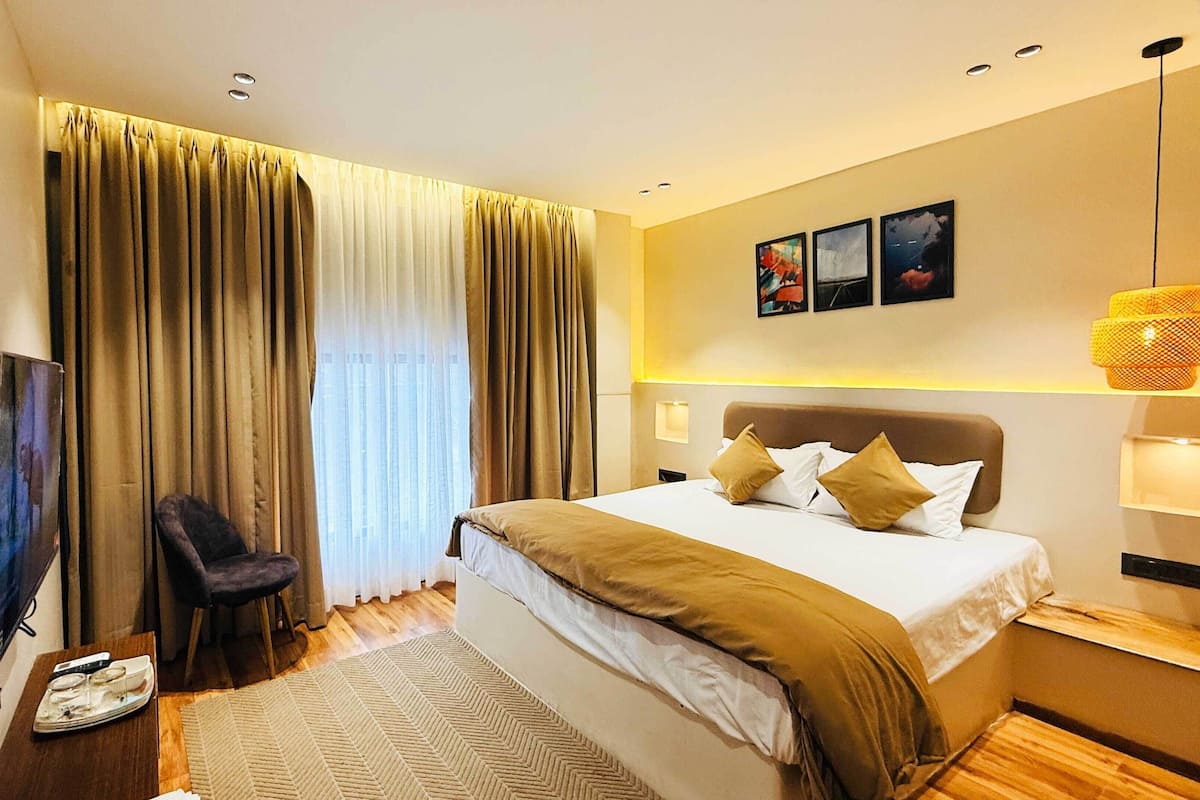 A modern hotel room with a large bed, a flat-screen TV, a chair, and a large window with curtains.