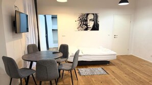 Una habitación moderna con una mesa redonda para comer, cuatro sillas grises, una cama con ropa de cama blanca, una obra de arte blanco y negro en la pared y un televisor montado.