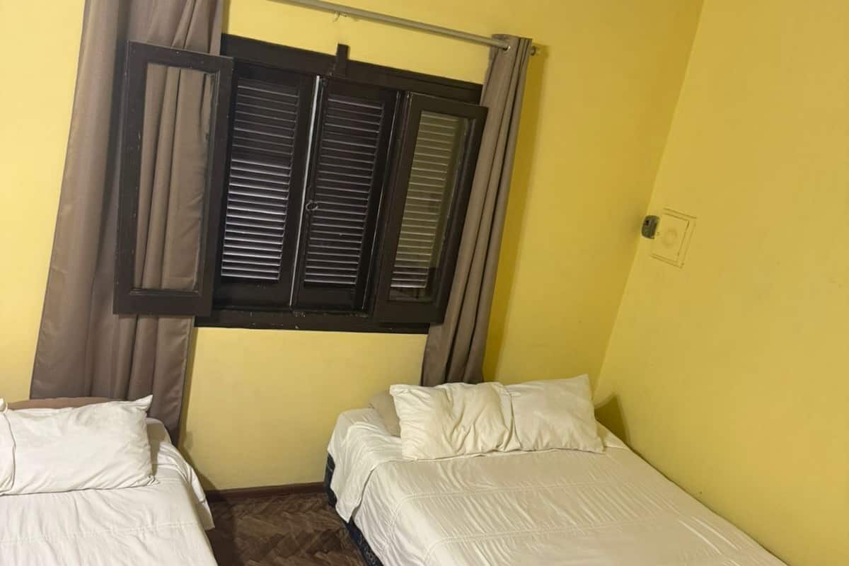 Una habitación con dos camas individuales, una ventana con cortinas marrones, un aire acondicionado instalado en el techo y un piso de madera con diseño de espiga.