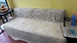 Un sofá con funda de ganchillo, una mesita con un florero y un periódico, y una ventana con vistas a la calle.