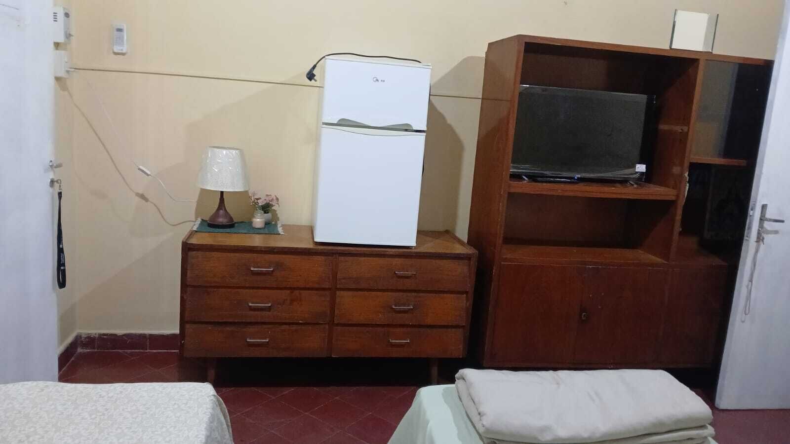 A small room with a wooden dresser, a white refrigerator, a television on a wooden stand, a bed with a white cover, and a small table with a lamp and flowers.