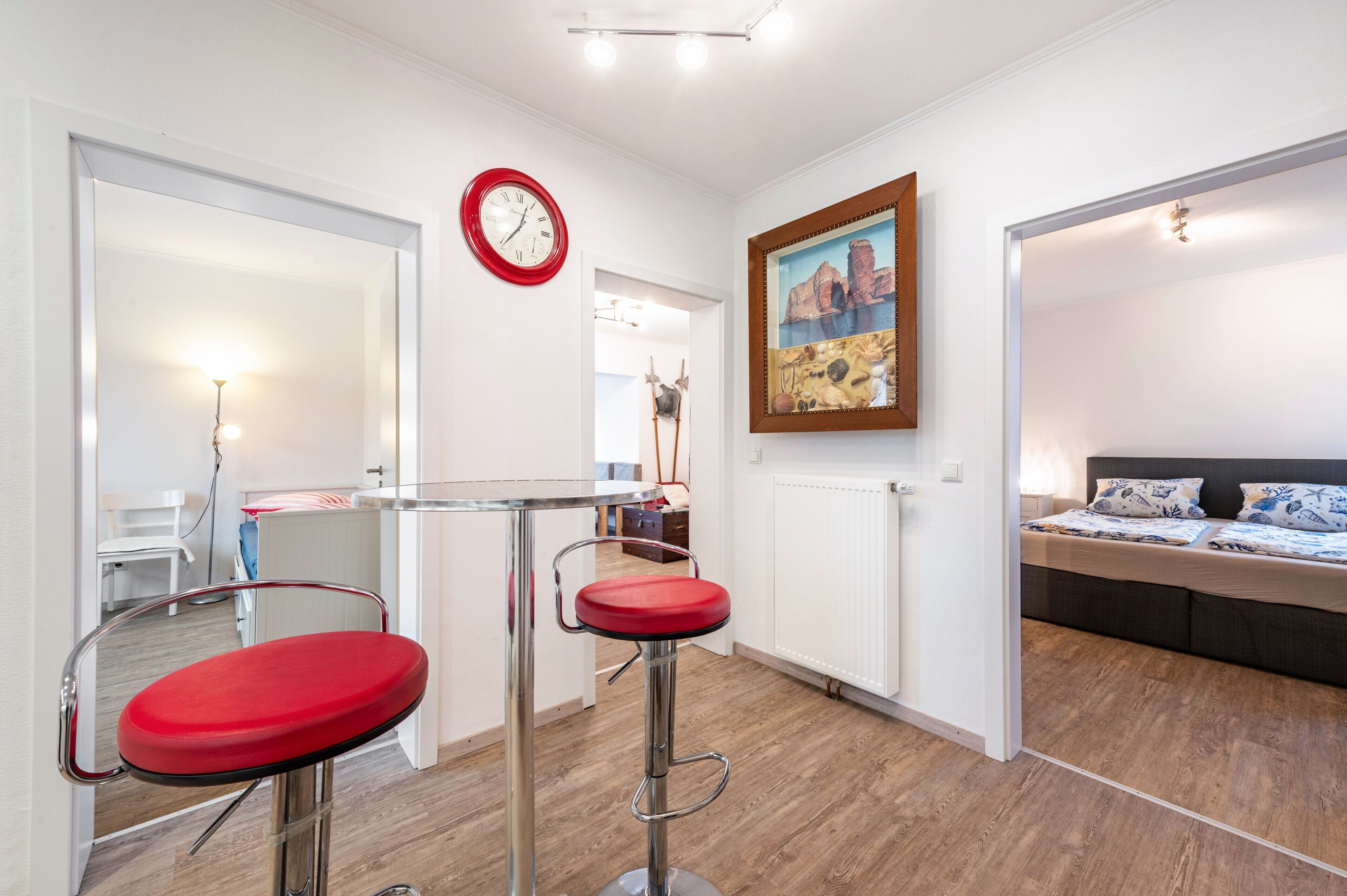 A room with a bed, a red clock, a painting, and a stool.