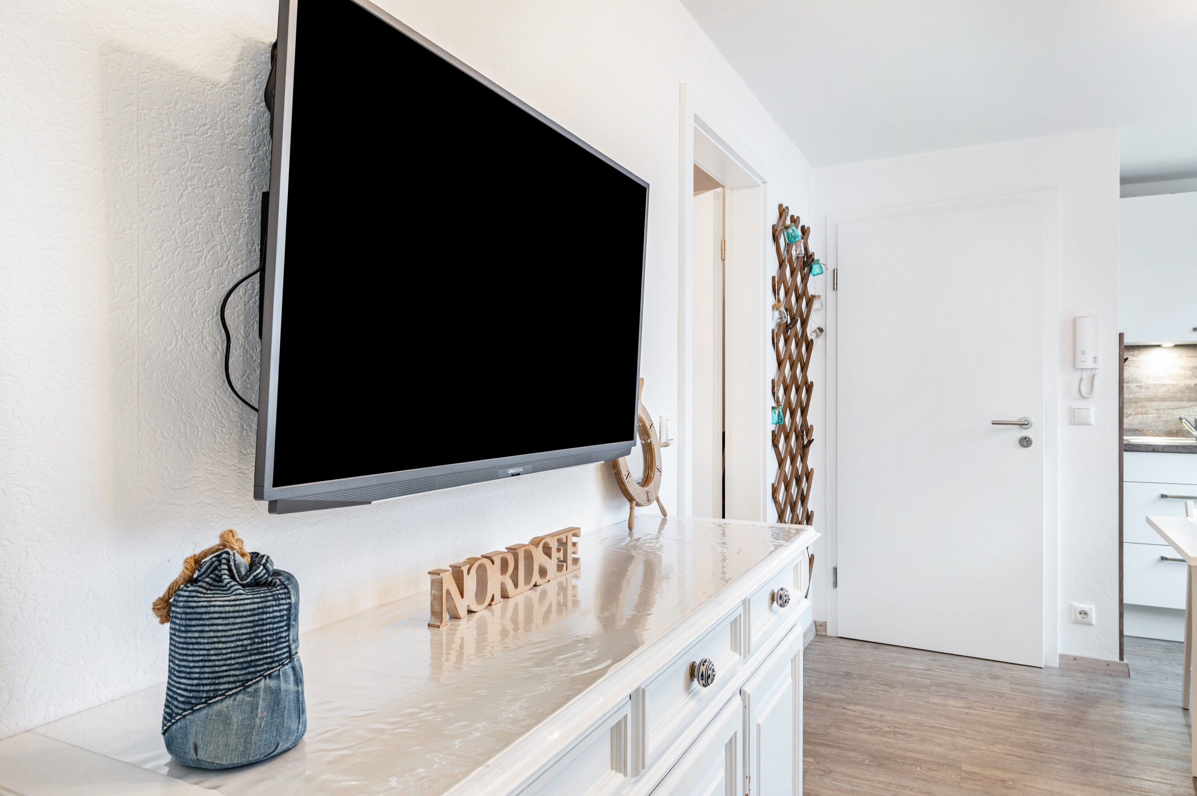 A modern living room with a flat-screen TV mounted on the wall, a wooden sign with letters, a small denim bag, and a decorative wall piece.