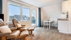 Ein modernes Wohnzimmer mit einer Sofagarnitur, einem Esstisch mit Stühlen, einer Küchenzeile und großen Fenstern.
