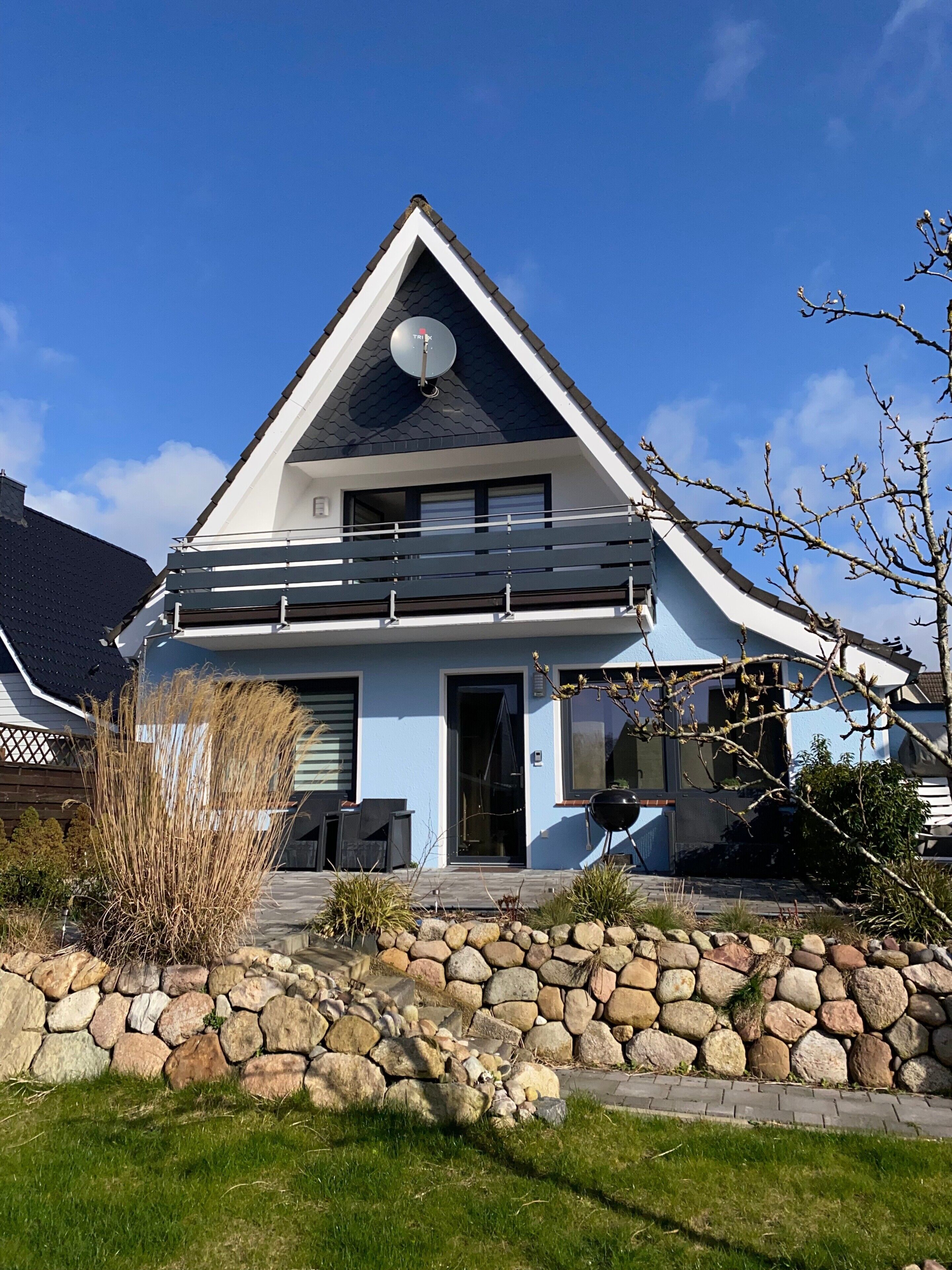 Une maison moderne à deux étages, avec un mur de pierre à l’avant, une antenne parabolique sur le toit et un ciel bleu dégagé.