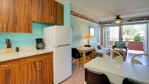 Cozinha compacta com armários de madeira, geladeira branca e cafeteira. Possui área de jantar com mesa e cadeiras, além de uma janela com cortinas.