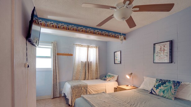 Quarto com duas camas, um ventilador de teto, janela com cortinas e um quadro na parede.